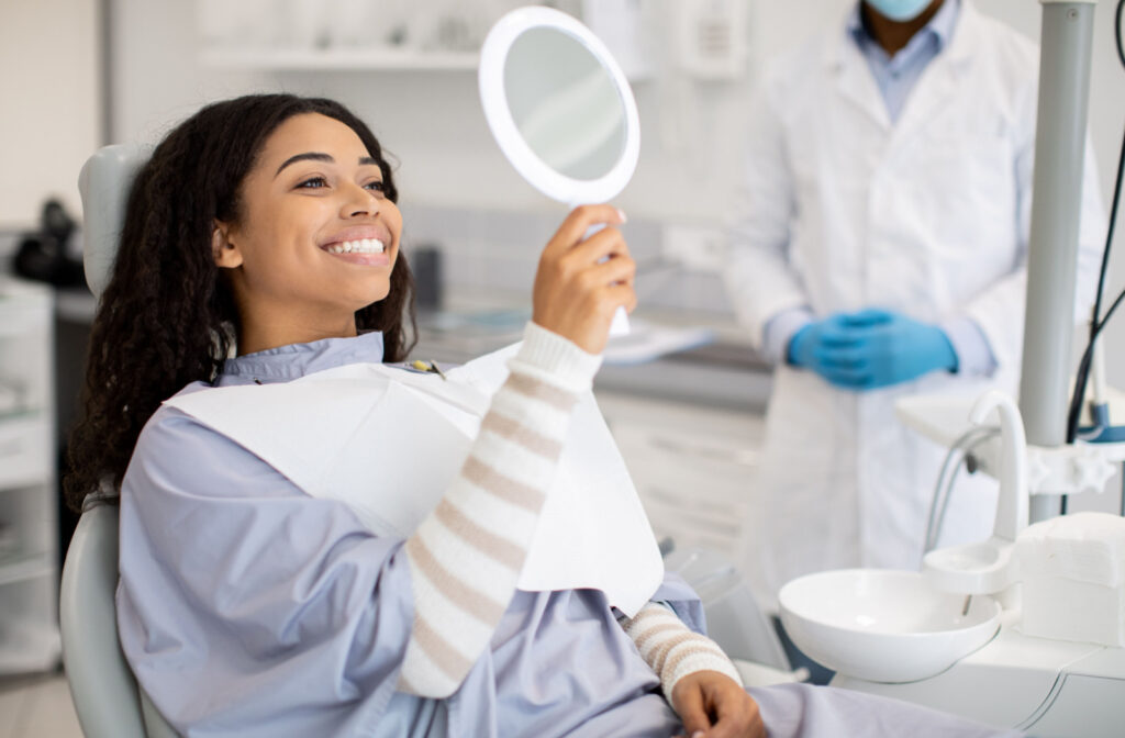 A woman sitting in a dentist's chair, holding up a mirror and smiling with her dentist in the background.
