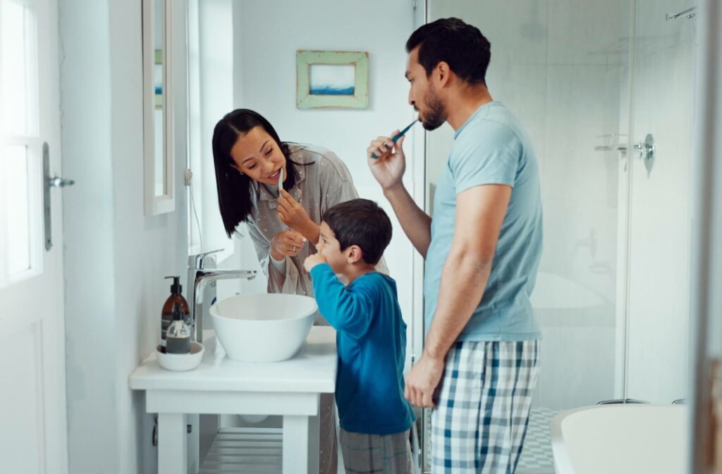 Parents teach their child how to brush their teeth together in a clean, white bathroom