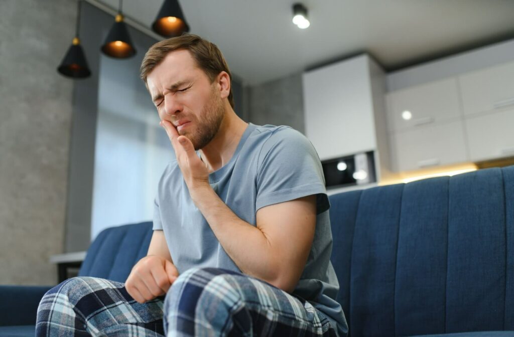 A man sitting on a couch and holding his right cheek in pain as he winces due to his teeth hurting.