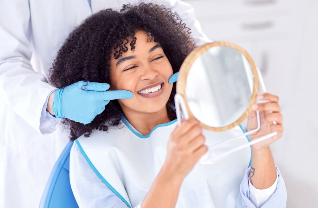 A patient smiling in a handheld mirror while an out-of-frame gloved dentist points to her new dental crown.