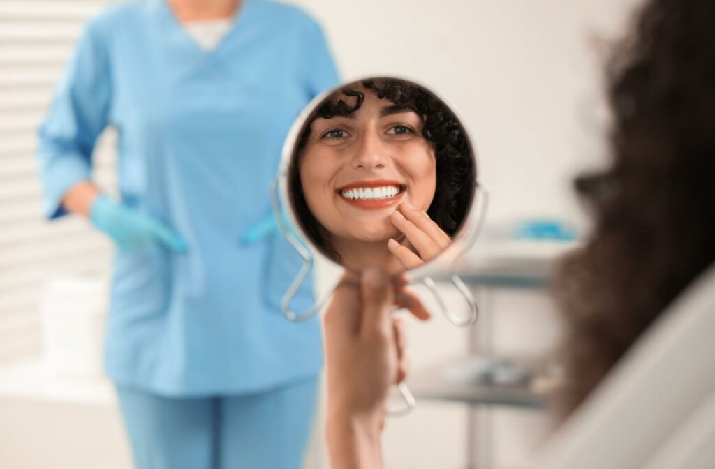 Person smiling in a mirror after a dental crown procedure with a dental professional visible in the background