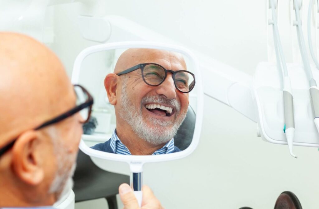 Smiling older man with glasses looking at his reflection in a handheld mirror showing off his new dental implants.