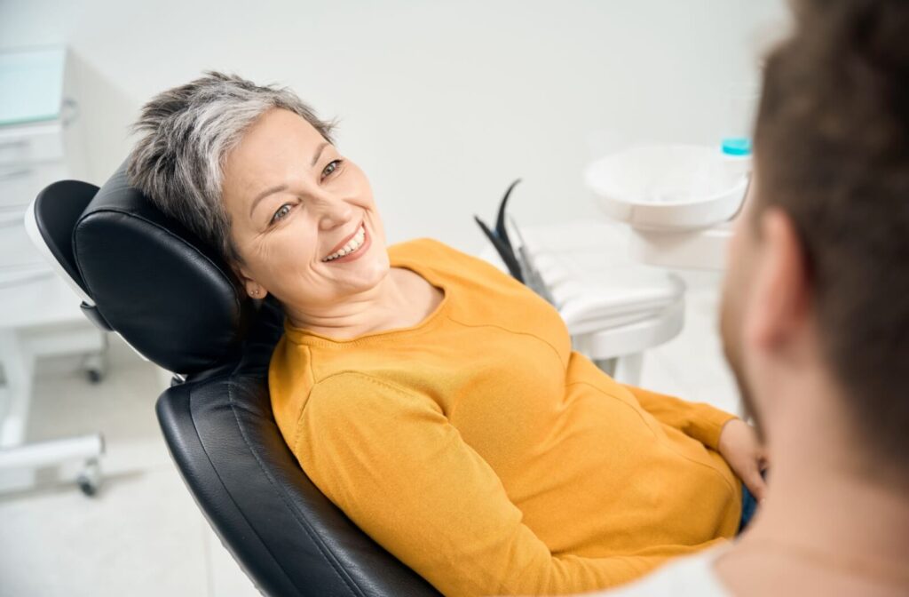 A young adult admires their newly whitened teeth in a hand mirror while their dentist stands behind them