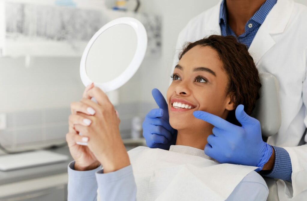 A young adult admires their newly whitened teeth in a hand mirror while their dentist stands behind them