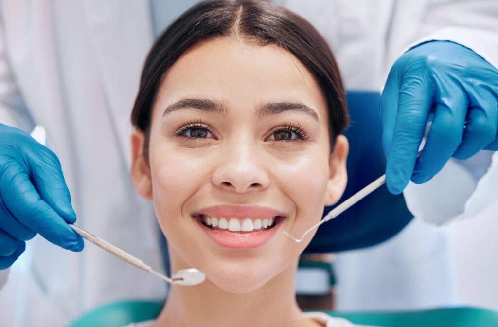A person smiling in a dental chair while a dentist wearing blue gloves holds dental tools near her teeth during a routine cleaning.